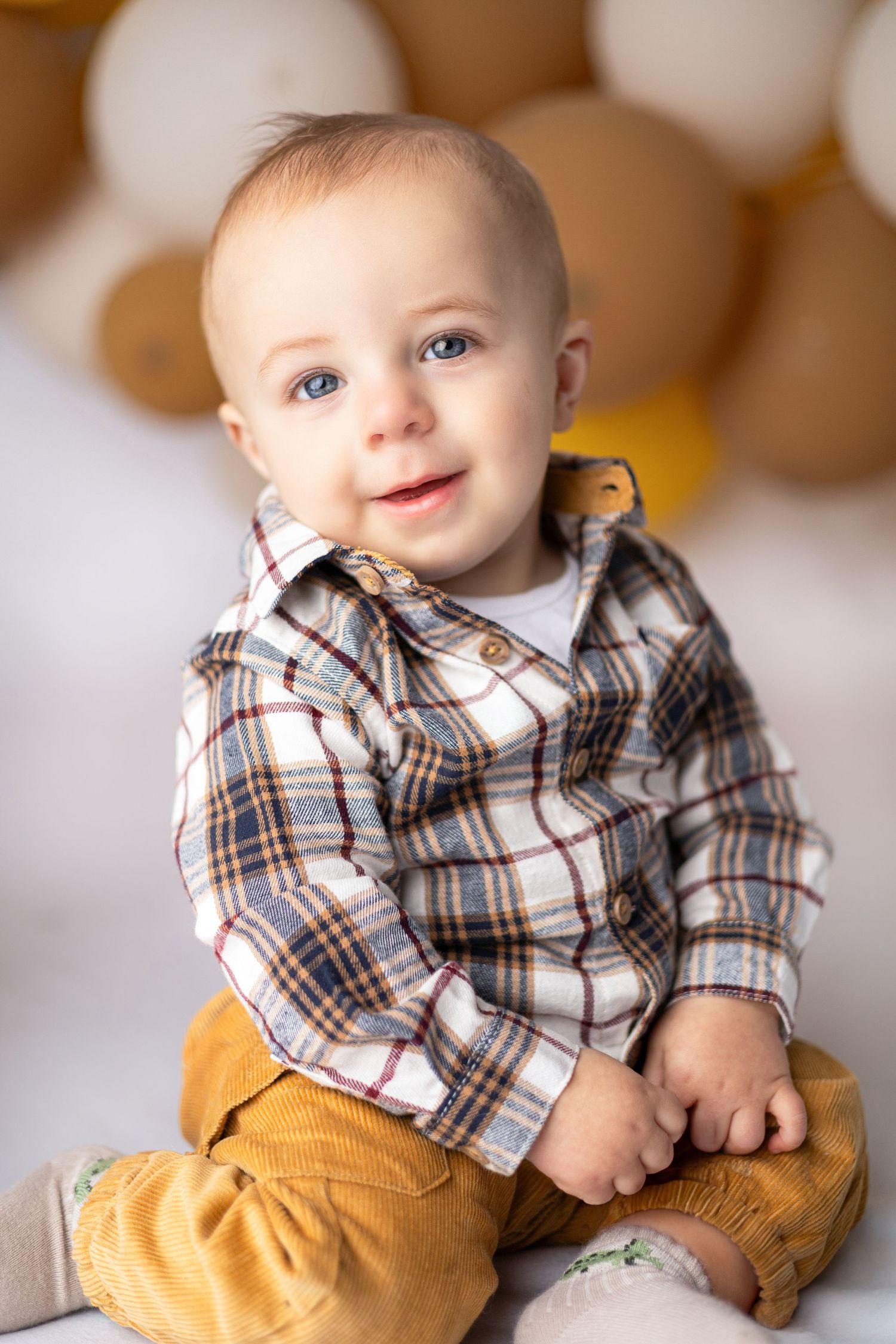 Baby wearing plaid shirt and mustard pants sitting against neutral background.