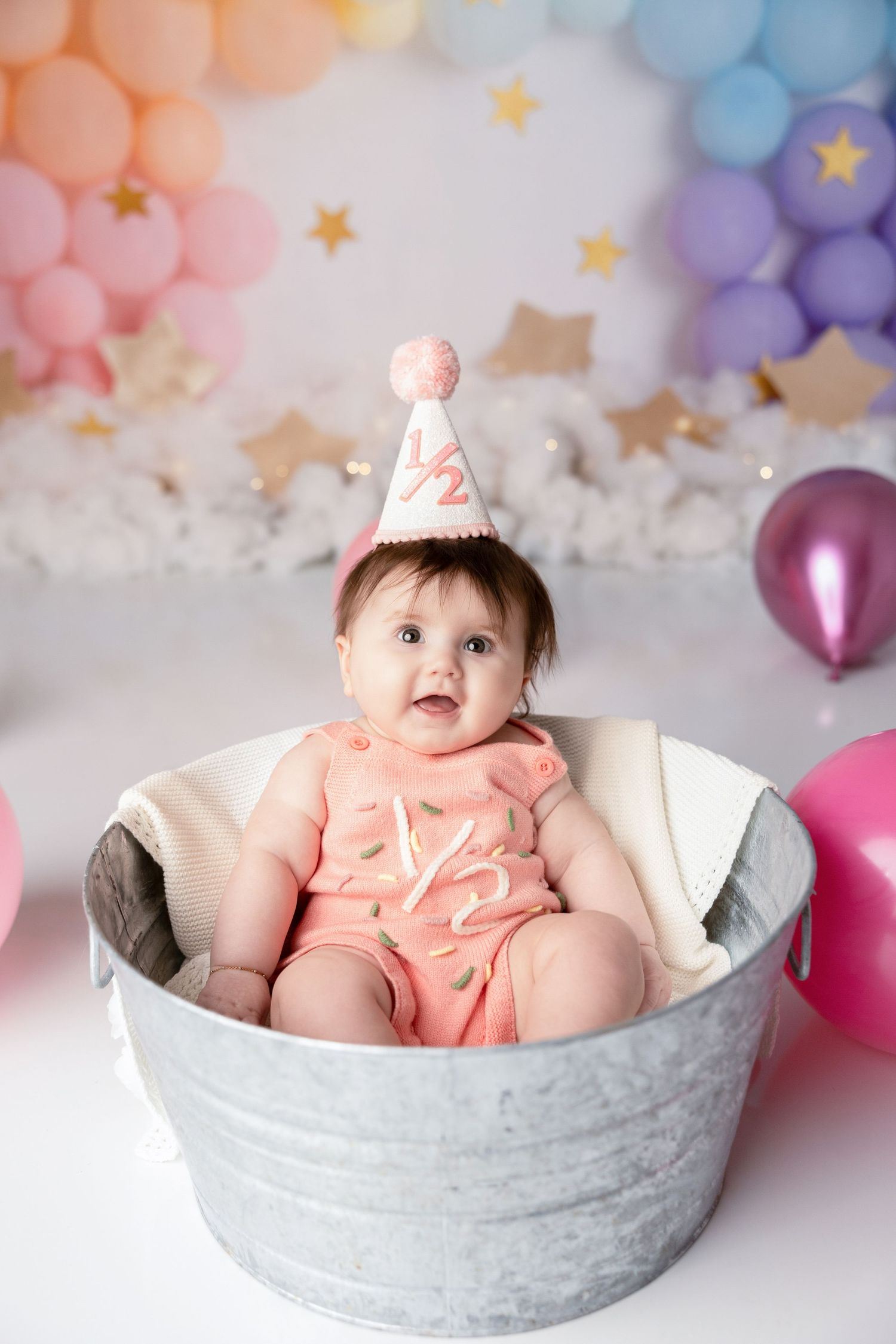 Baby sits in grey basket surrounded by pastel balloons and star decorations.
