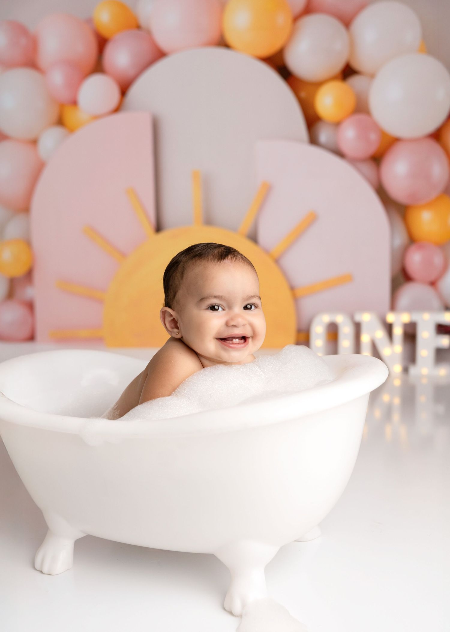 A baby sits in a white decorative bathtub with pink and orange balloons in the background.