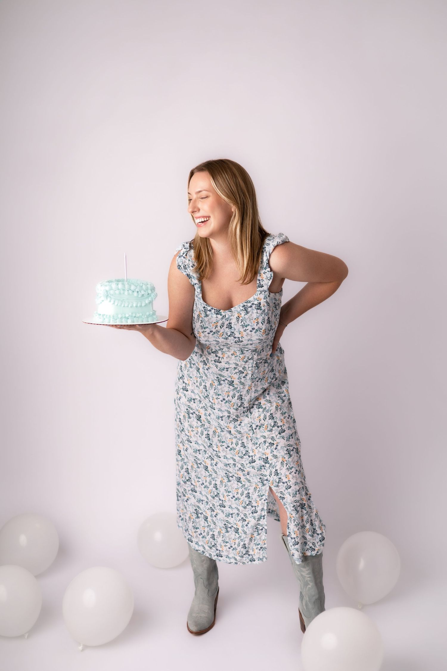 Woman in floral dress stands energetically with birthday cake surrounded by white balloons.