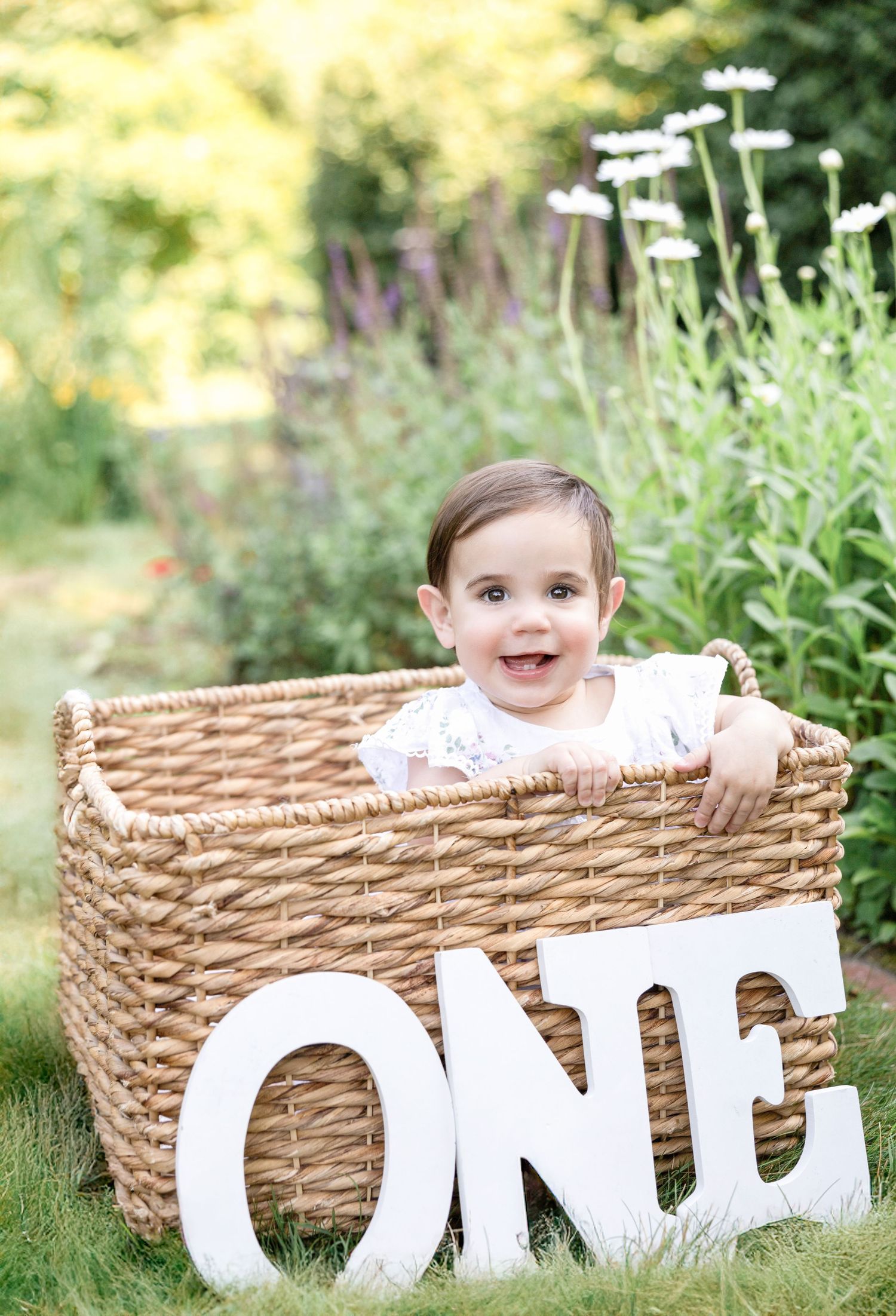A baby poses with white letter blocks spelling ONE in a woven basket outdoors.