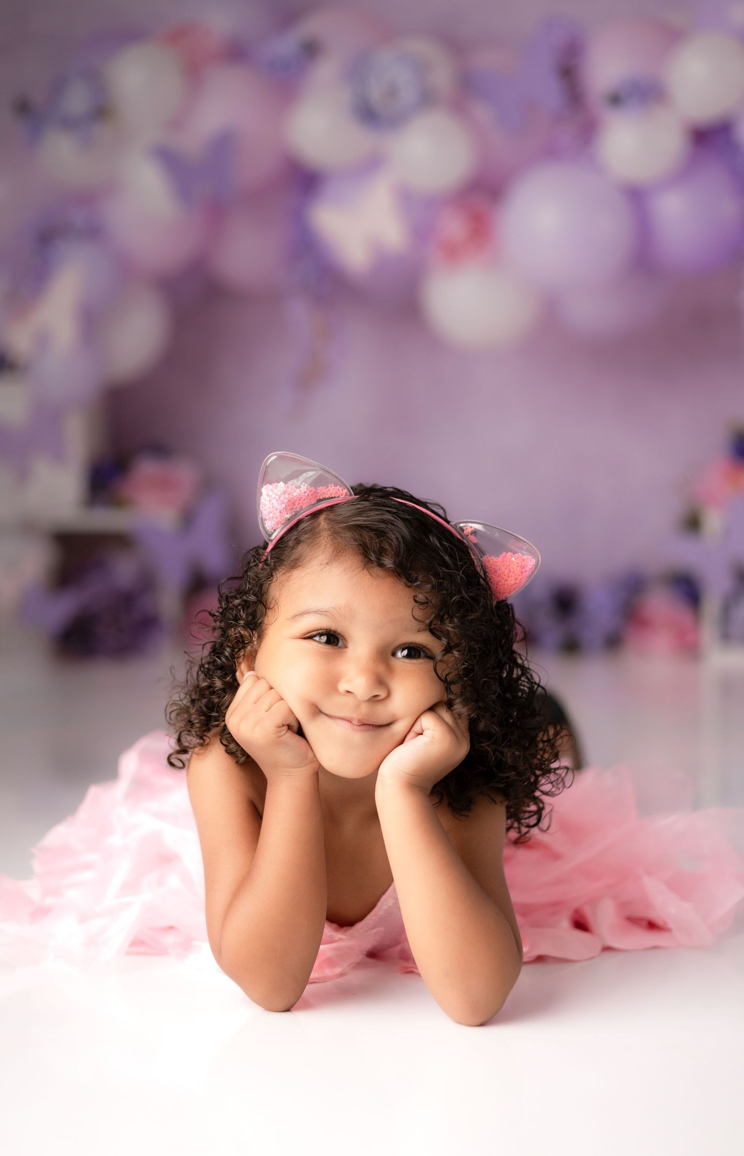 Child with curly hair posing playfully on white floor with purple bokeh lights.