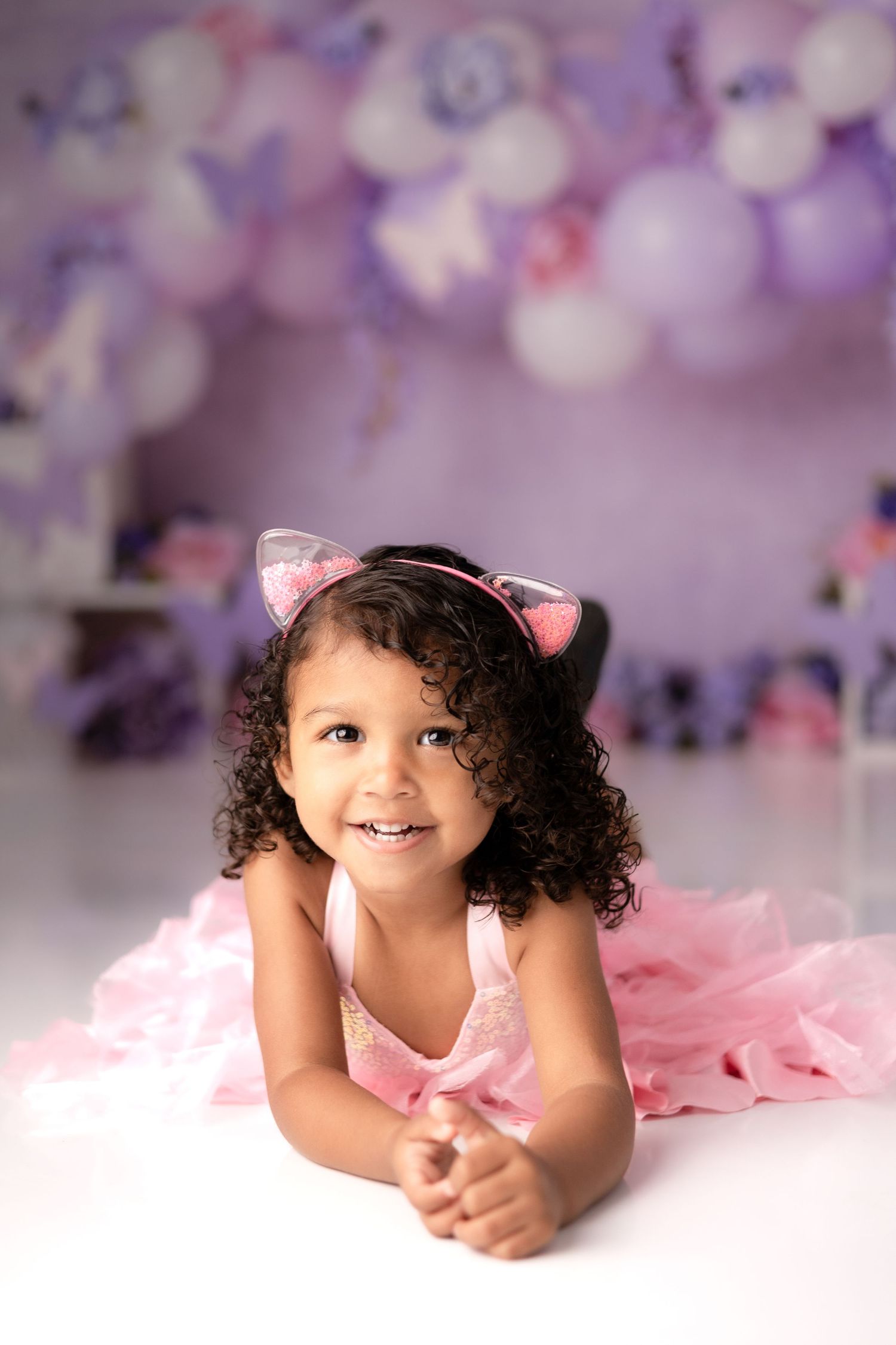 Young child in pink tulle lying on white floor with purple bokeh background in studio setting.