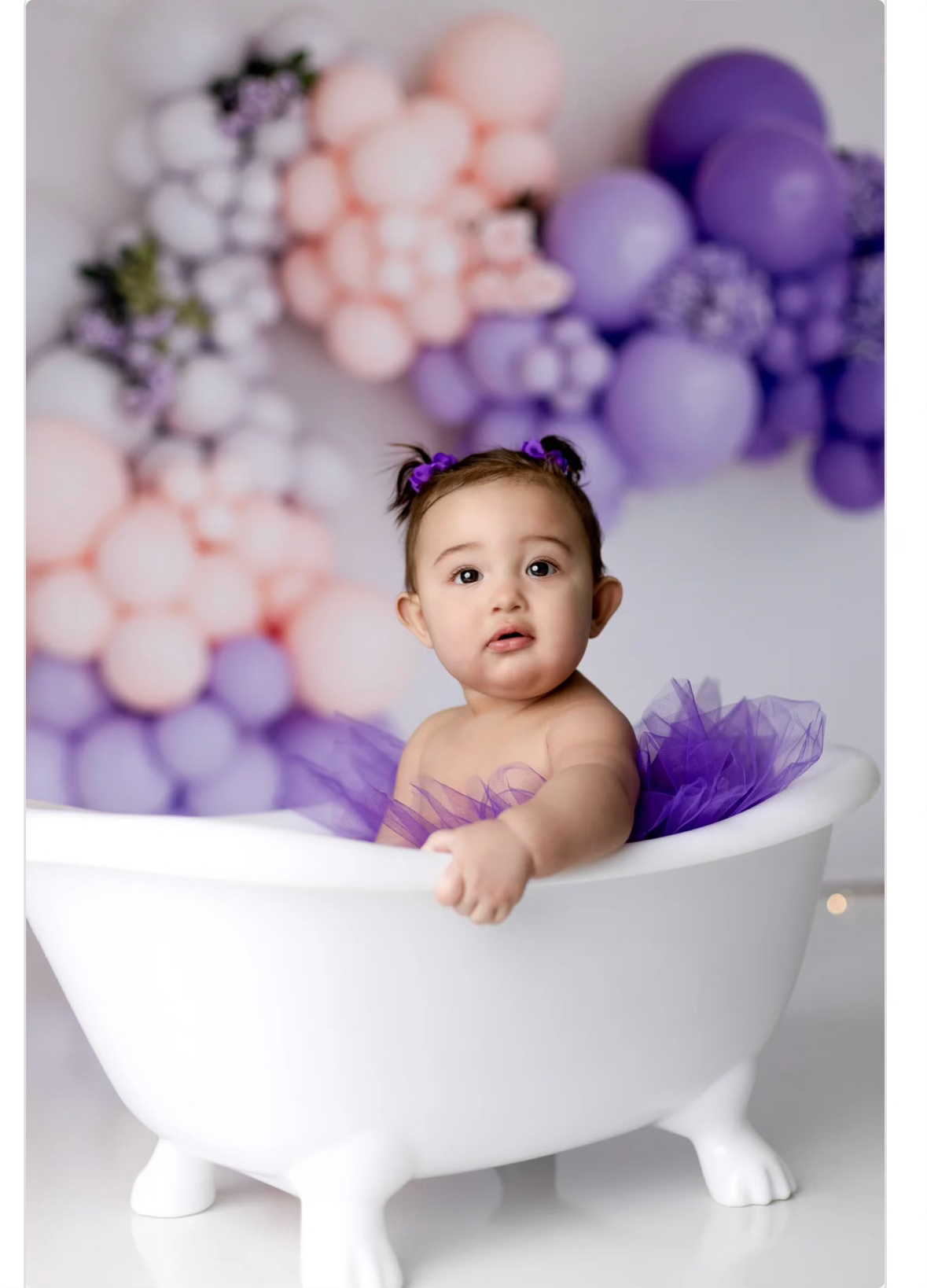 Baby in purple tutu sits in decorative white bathtub with pink and purple balloon backdrop.