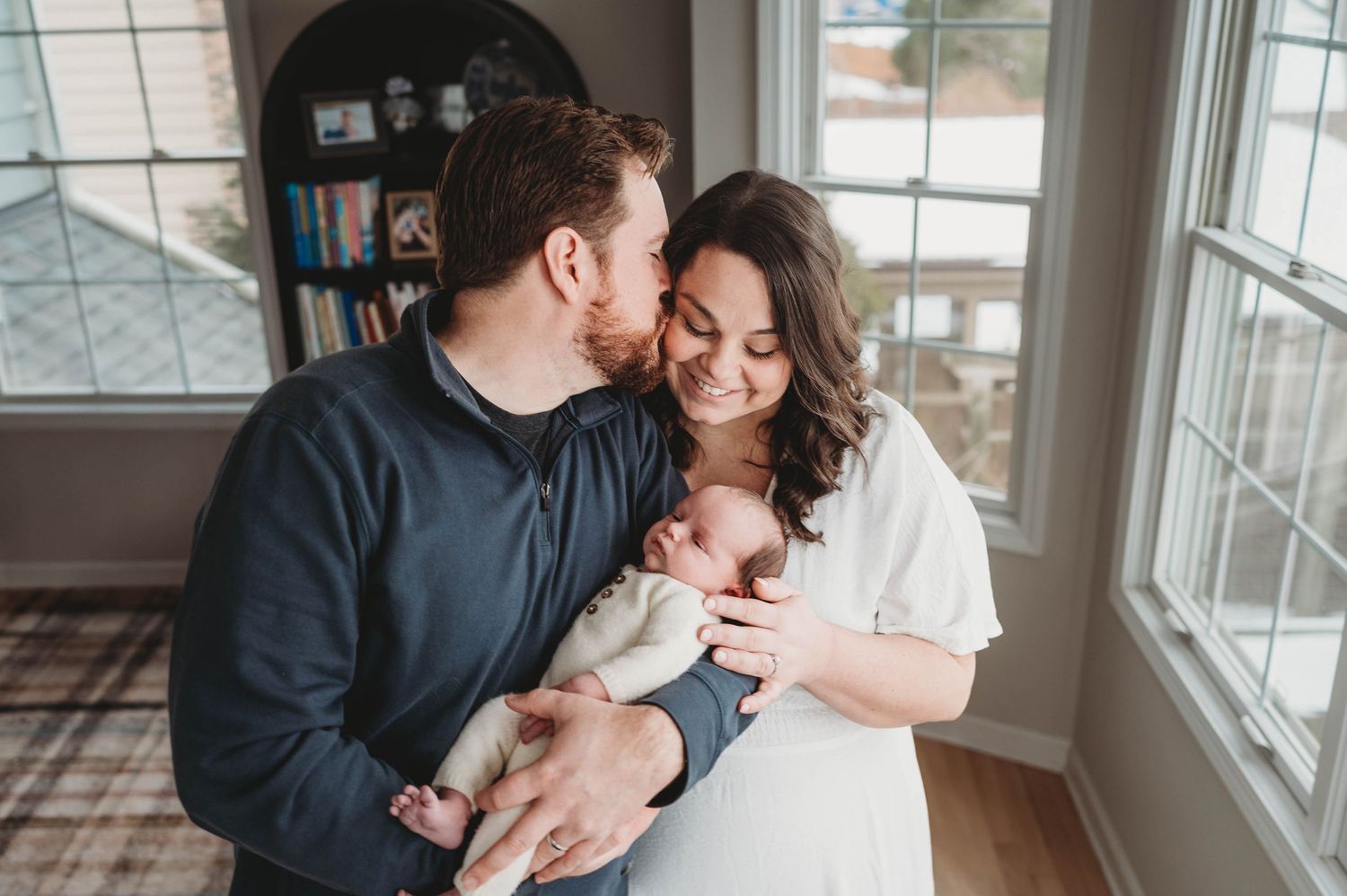 Couple sharing a tender kiss moment while holding their sleeping newborn baby by a bright window.