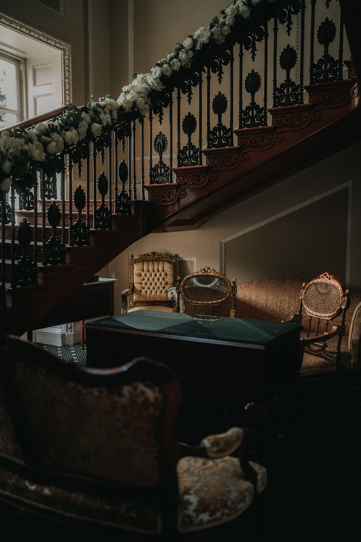 A dimly lit vintage staircase decorated with garland and antique chairs at the bottom creating an elegant atmosphere.
