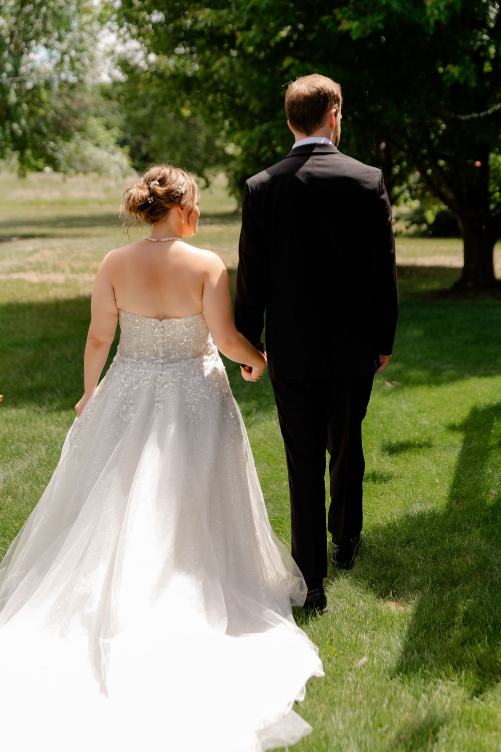 Bride in white beaded gown and groom in black tuxedo walking hand in hand through lush green garden.