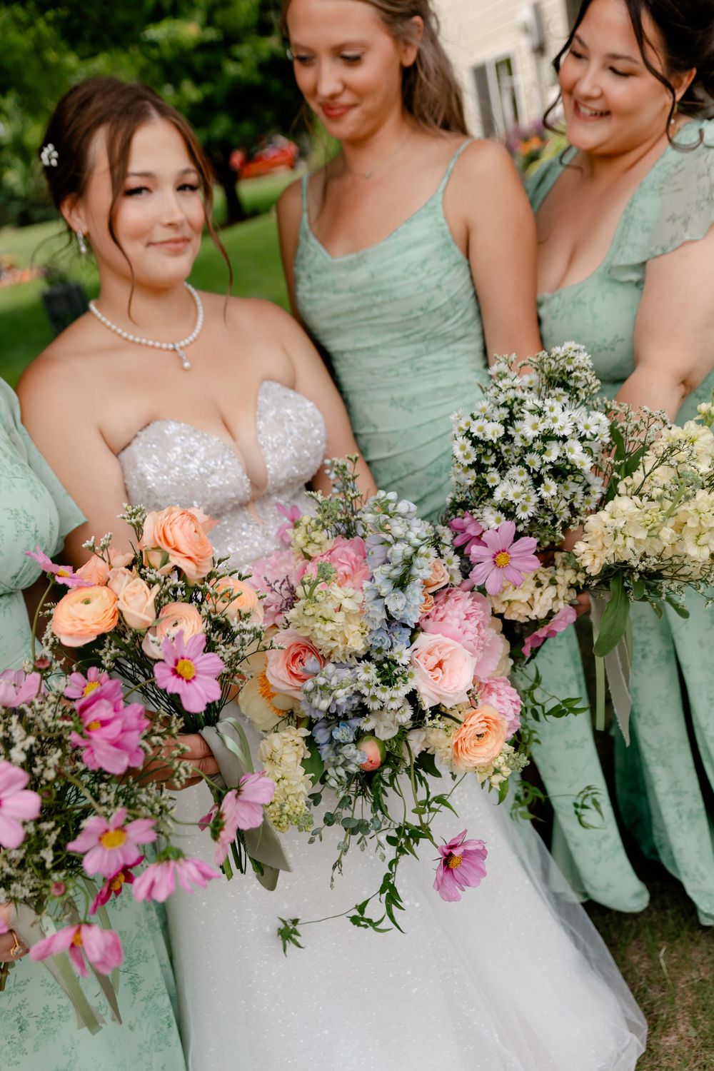 Bride in white strapless gown holds colorful bouquet with bridesmaids in mint green dresses at outdoor wedding.