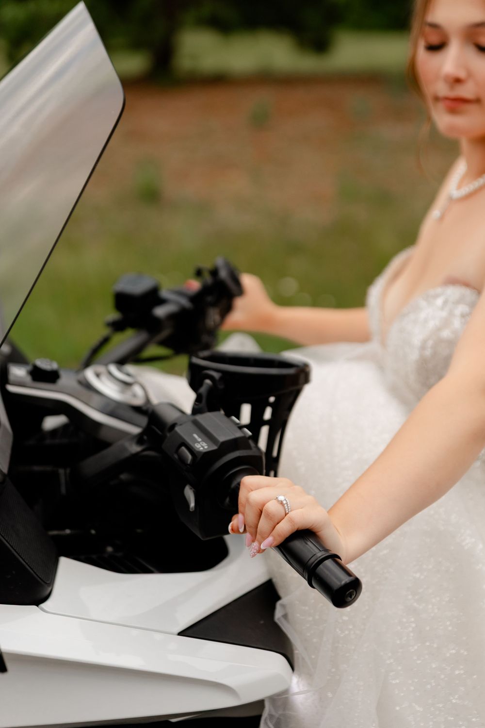 Bride in a sparkly white wedding gown holding the handlebars of a white motorcycle outdoors.