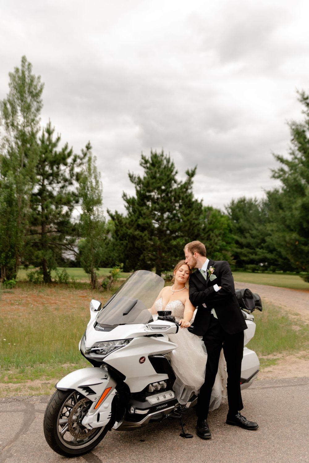 Bride and groom posing with a white Honda Gold Wing motorcycle on a cloudy day with green trees in the background.
