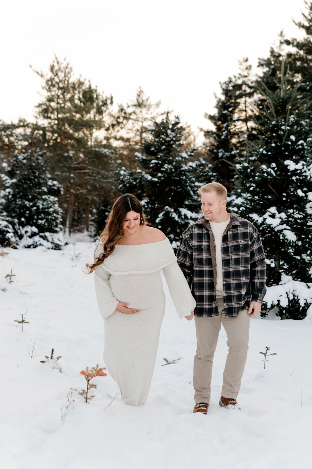 Pregnant couple holding hands in snowy winter forest, woman in white off-shoulder maternity dress, man in plaid shirt.