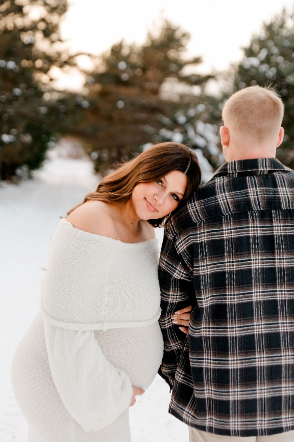 Smiling pregnant woman in white knit dress leaning against partner in snowy winter setting.