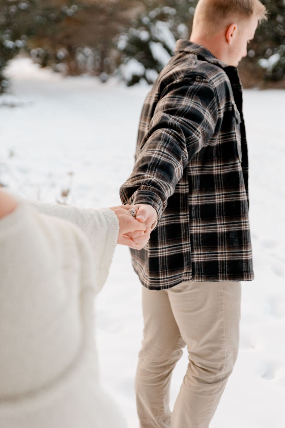 Couple holding hands walking through snow, man in black plaid flannel shirt and khaki pants.