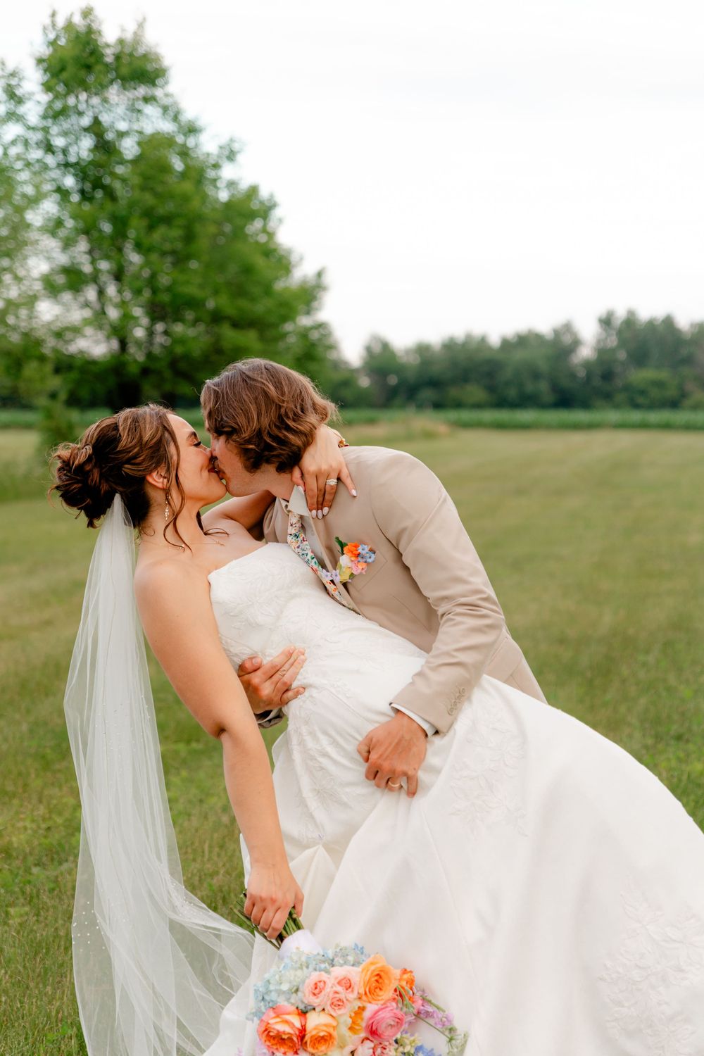 A bride and groom share a romantic dip kiss outdoors on green grass, she holds a colorful bouquet in a white gown.
