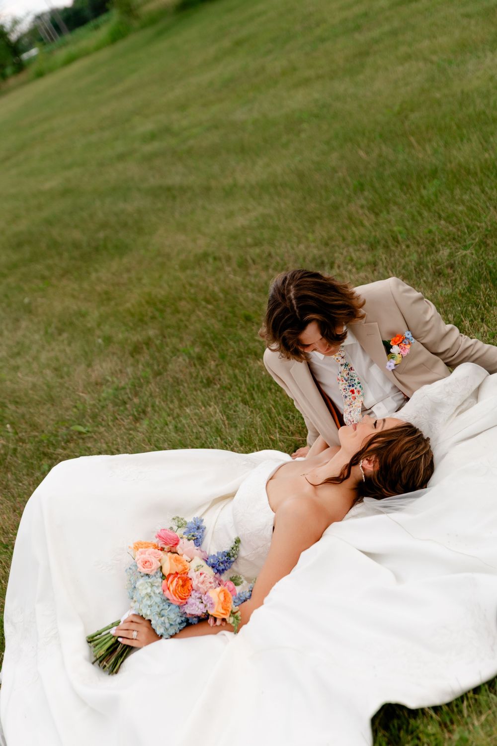 Bride lying in grass holding colorful bouquet while groom leans over her during romantic outdoor wedding photo session.