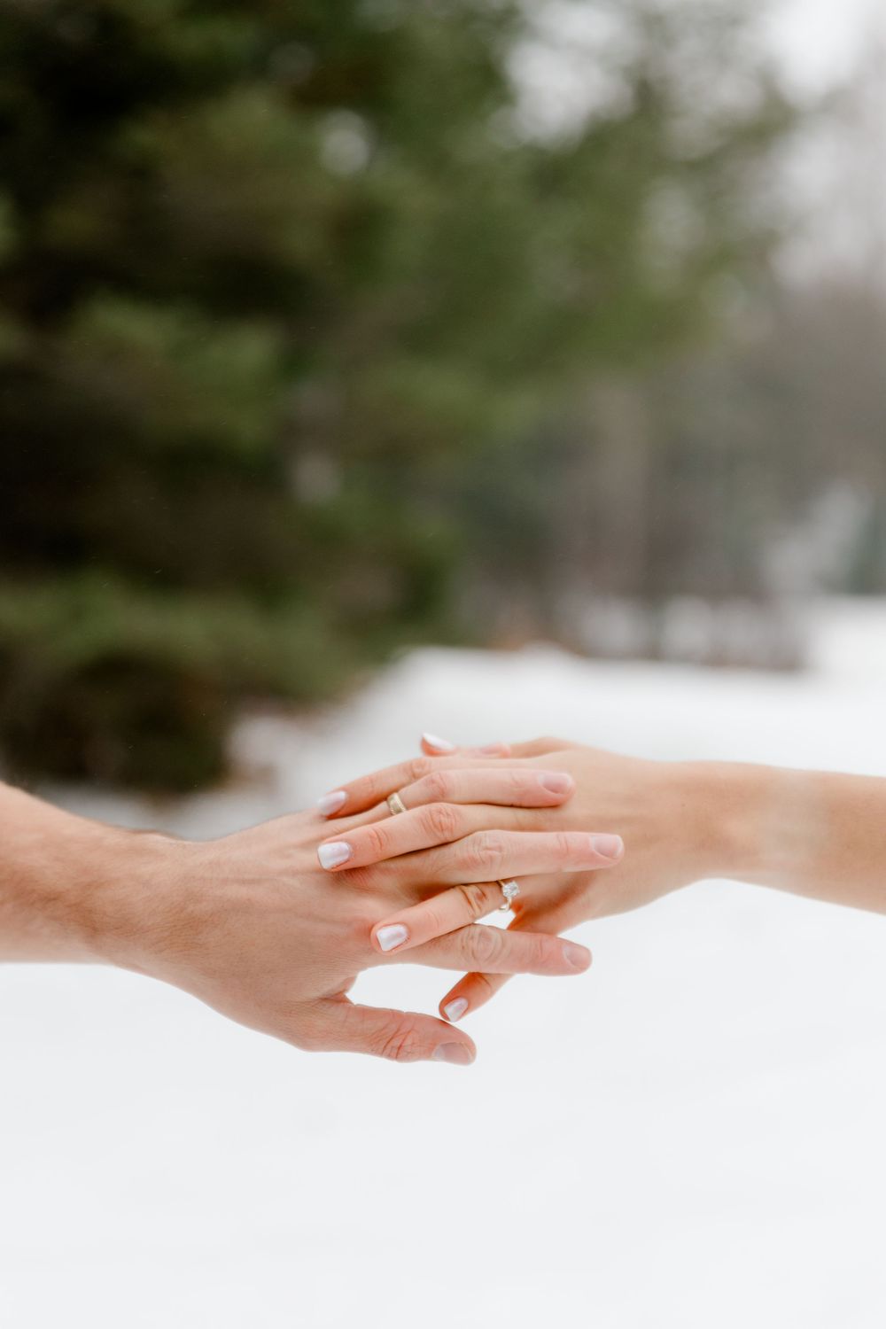 Close-up of an engaged couple holding hands showing gold engagement rings against a snowy background.