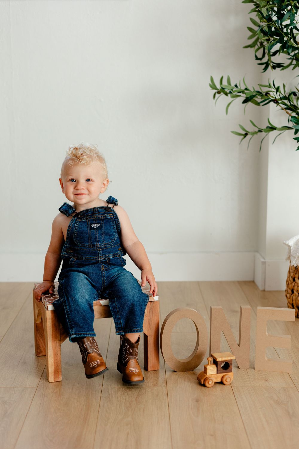 Toddler boy in denim overalls and boots sits on wooden block beside ONE letters for first birthday photo.