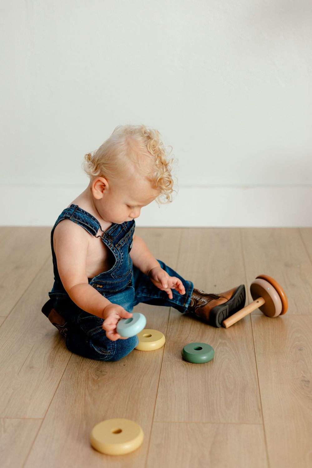 Curly-haired toddler in denim overalls sitting on wooden floor playing with colorful wooden stacking ring toy.