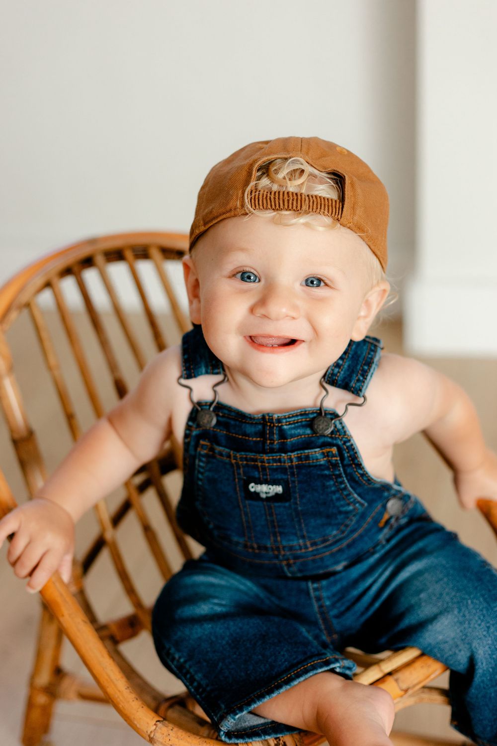Smiling baby in denim OshKosh overalls and brown cap sits in a wicker rocking chair.