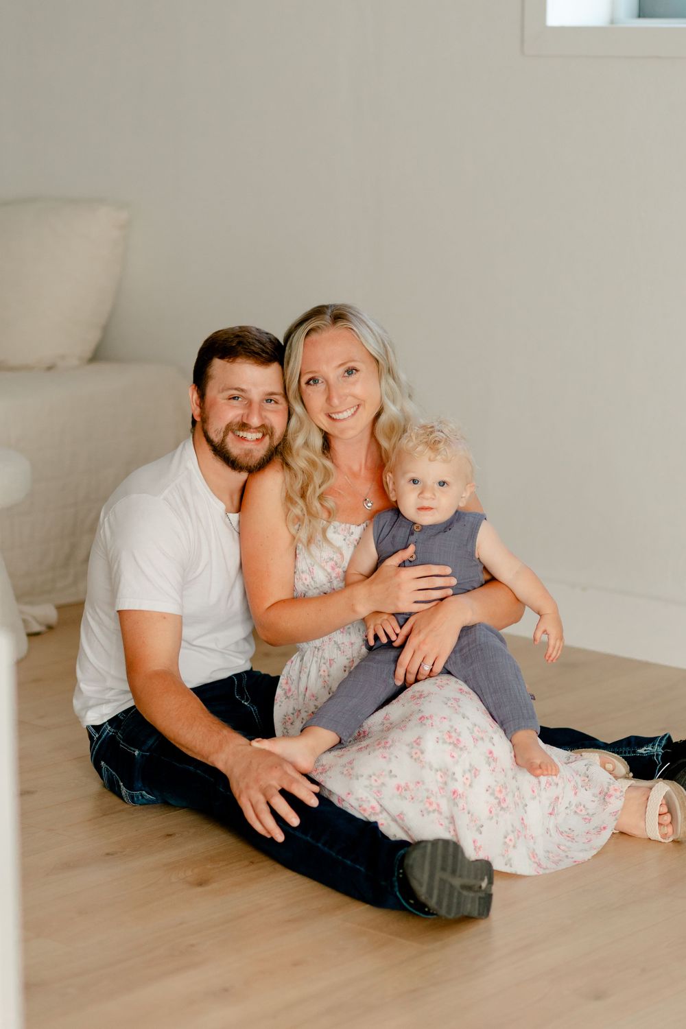 A smiling young family with a man, woman, and baby sitting together on a light wood floor in a bright white room.