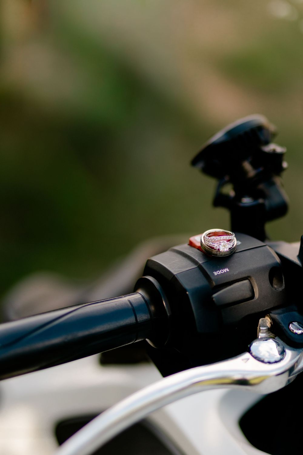 Close-up of a diamond engagement ring placed on a motorcycle handlebar control switch, with blurred green background.