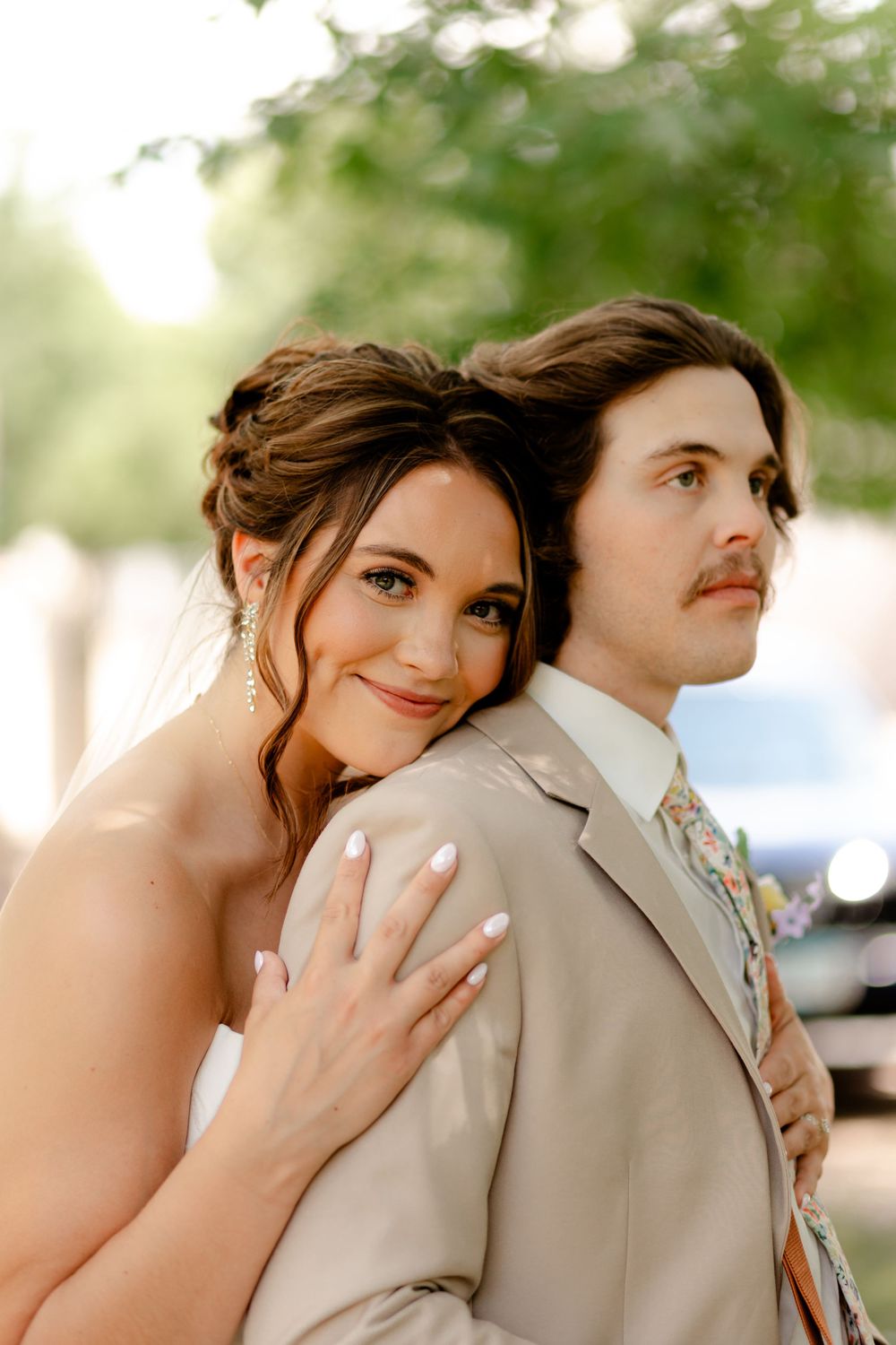 Bride in white dress embraces groom in tan suit with floral tie, posing outdoors with soft green bokeh background.