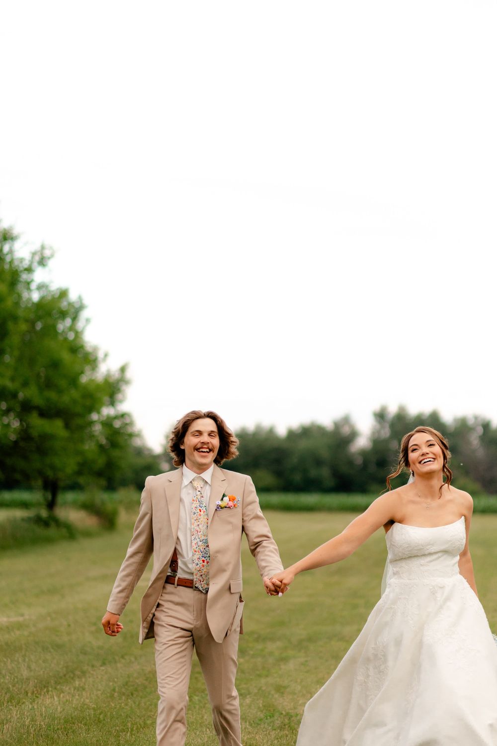 A joyful bride and groom hold hands while walking through a green field, the groom in a tan suit and bride in a white gown.