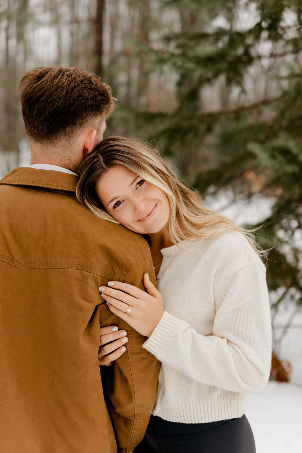 A woman in a cream sweater smiles at the camera while hugging a man in a brown jacket in a snowy forest.