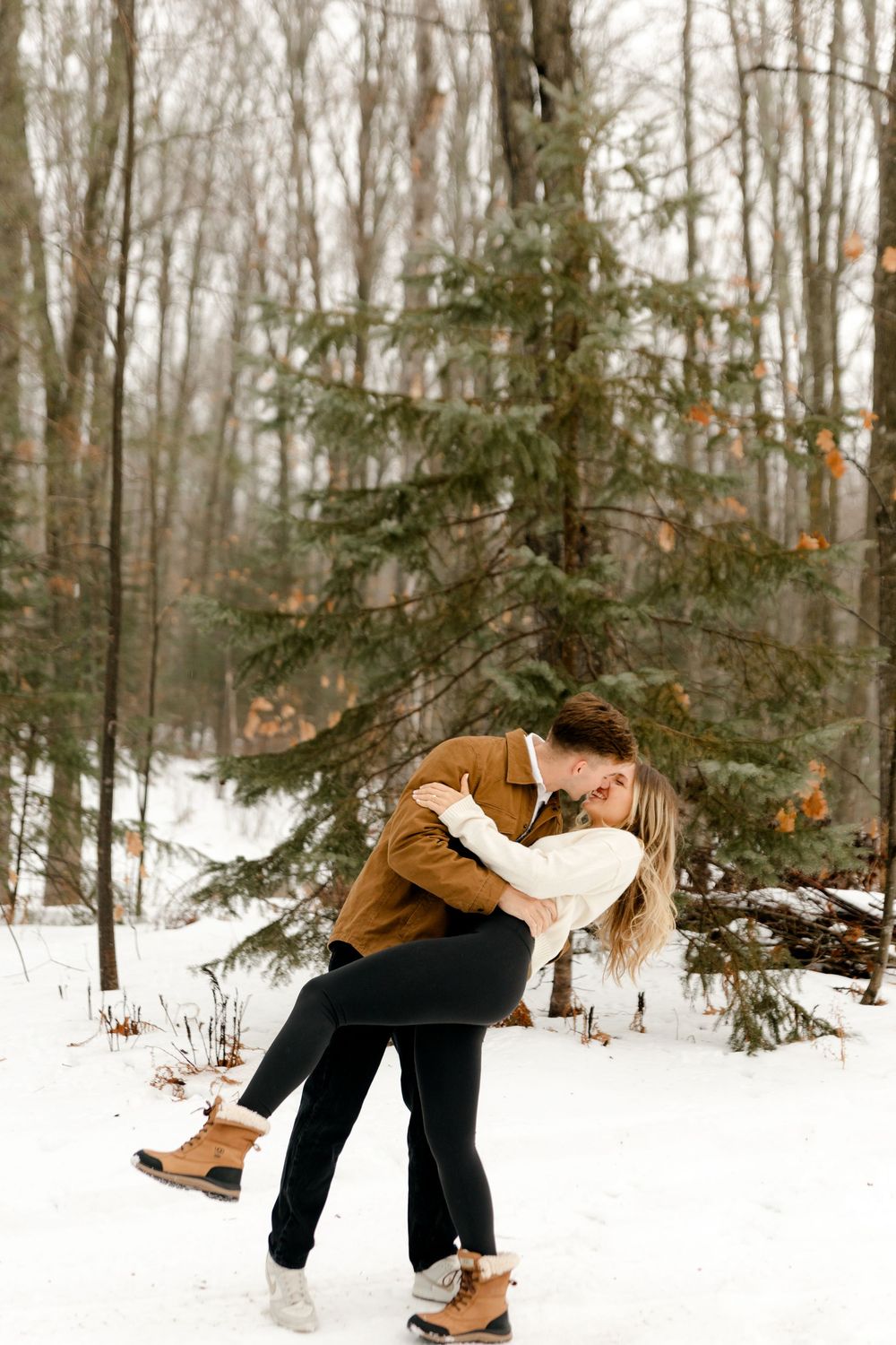 A couple shares a romantic dip kiss in a snow-covered forest with evergreen trees in the background.