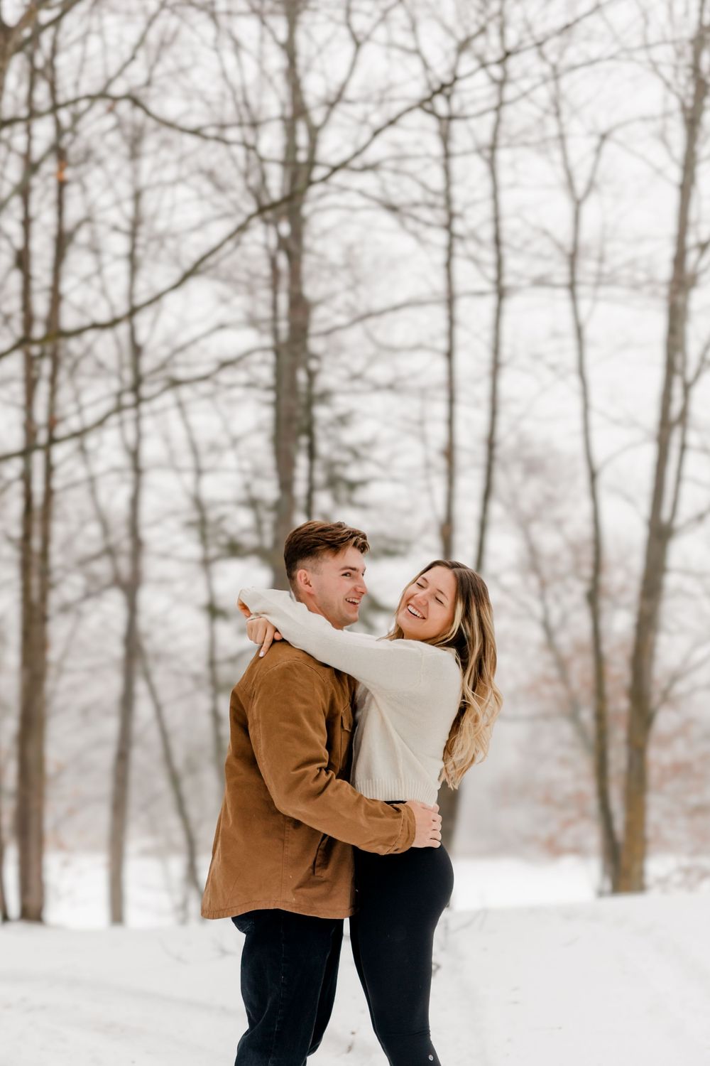 A laughing couple embraces in a snowy winter landscape surrounded by bare trees, wearing casual winter outfits.