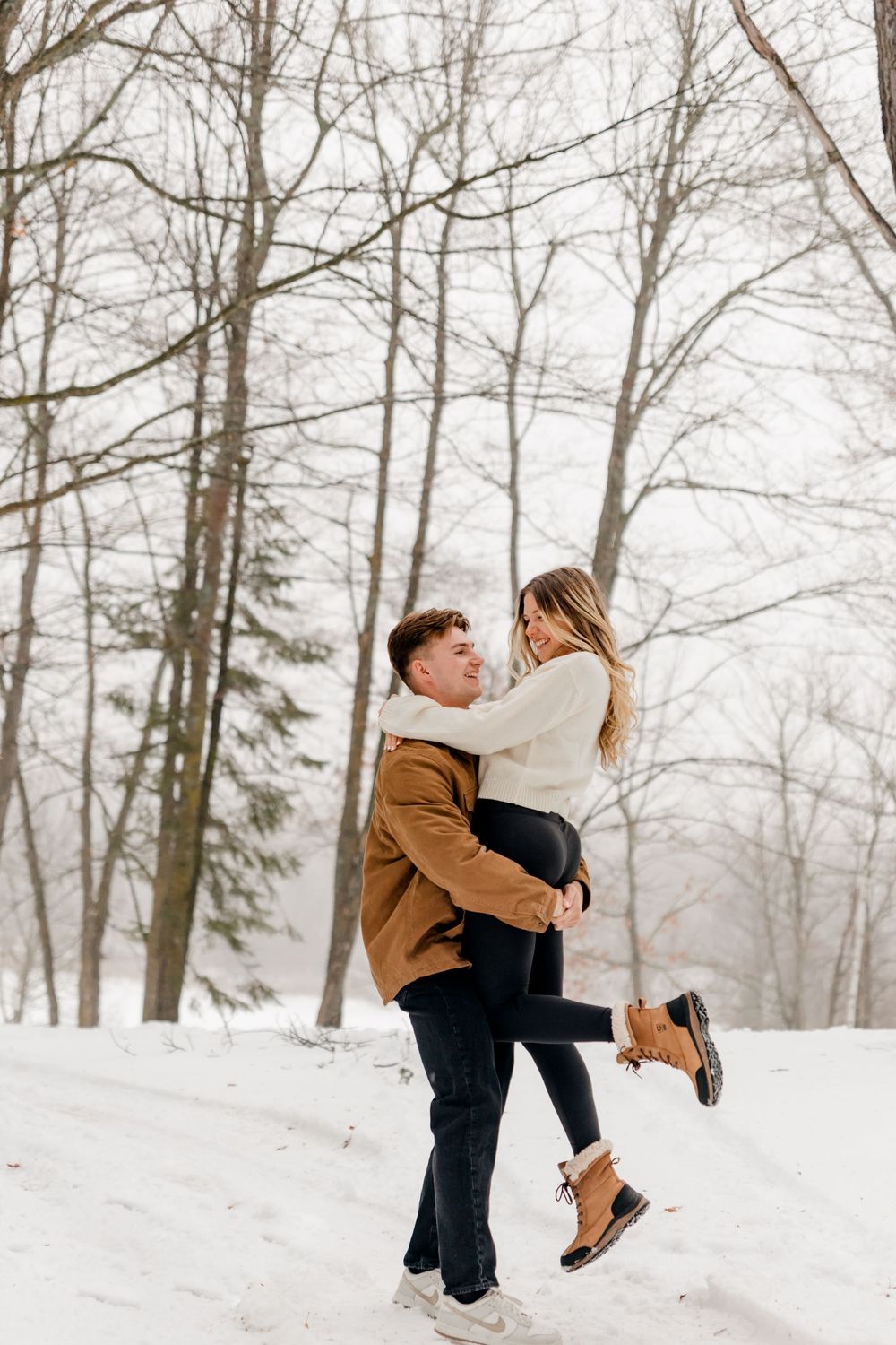 A man lifts a smiling woman in a cream sweater in a snowy winter forest during a couples photo session.