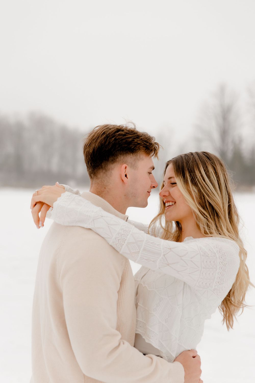 A joyful couple in matching cream sweaters nearly kisses while embracing in a bright snowy winter field.