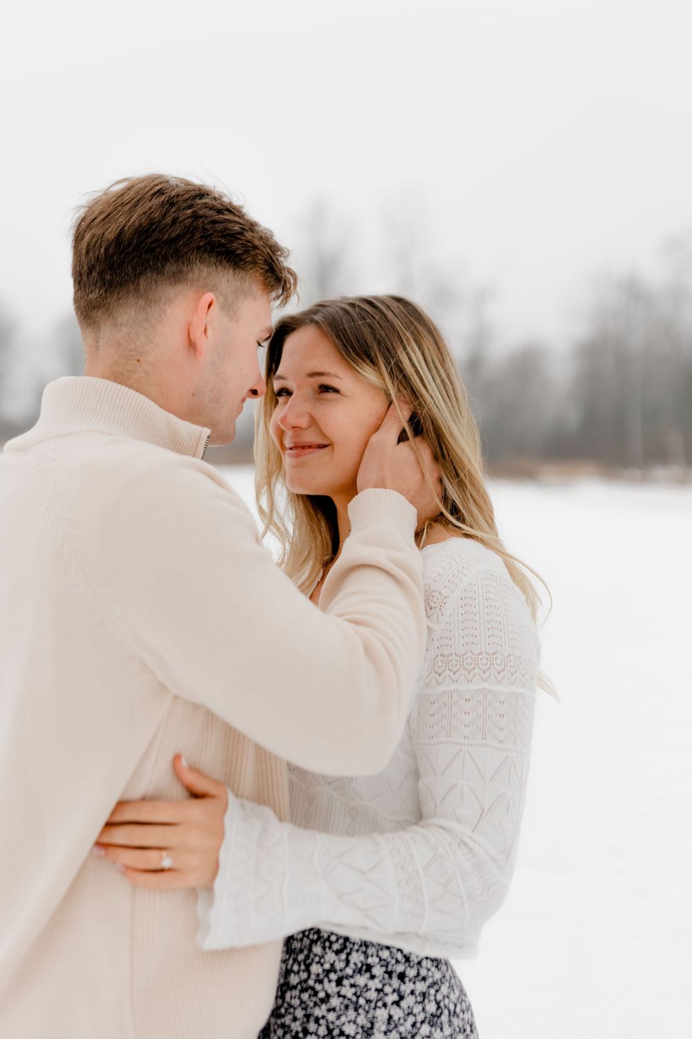 A man gently cups a smiling womans face as they embrace closely in a serene snowy winter setting.
