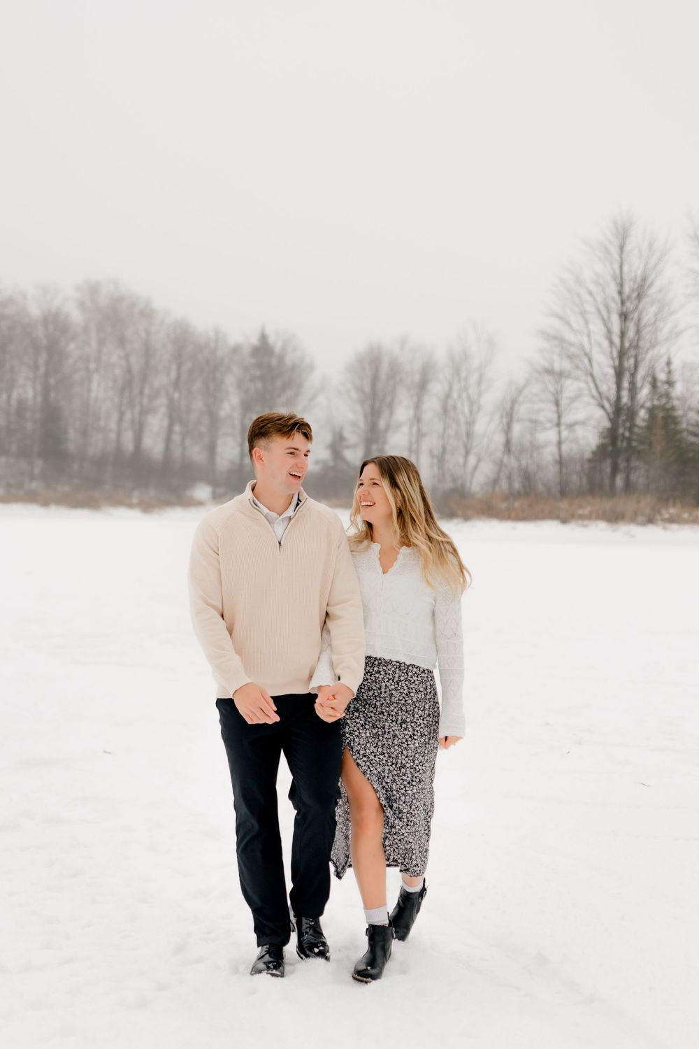 Happy couple walking hand in hand through a snowy field, smiling at each other in winter outfits.