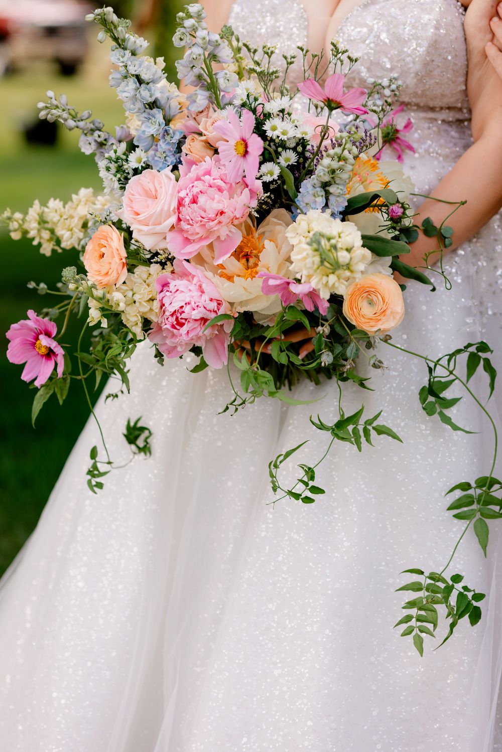 Bride holding a lush wedding bouquet with pink peonies, peach roses, blue delphinium, and trailing greenery against a white gown.