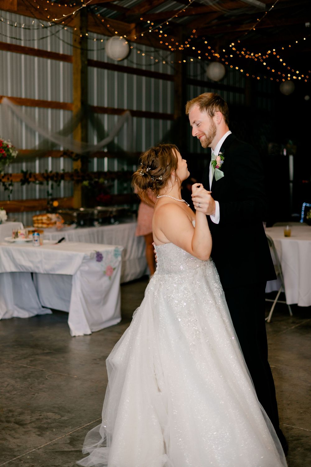 Bride and groom share their first dance in a rustic barn wedding venue decorated with string lights and white draping.
