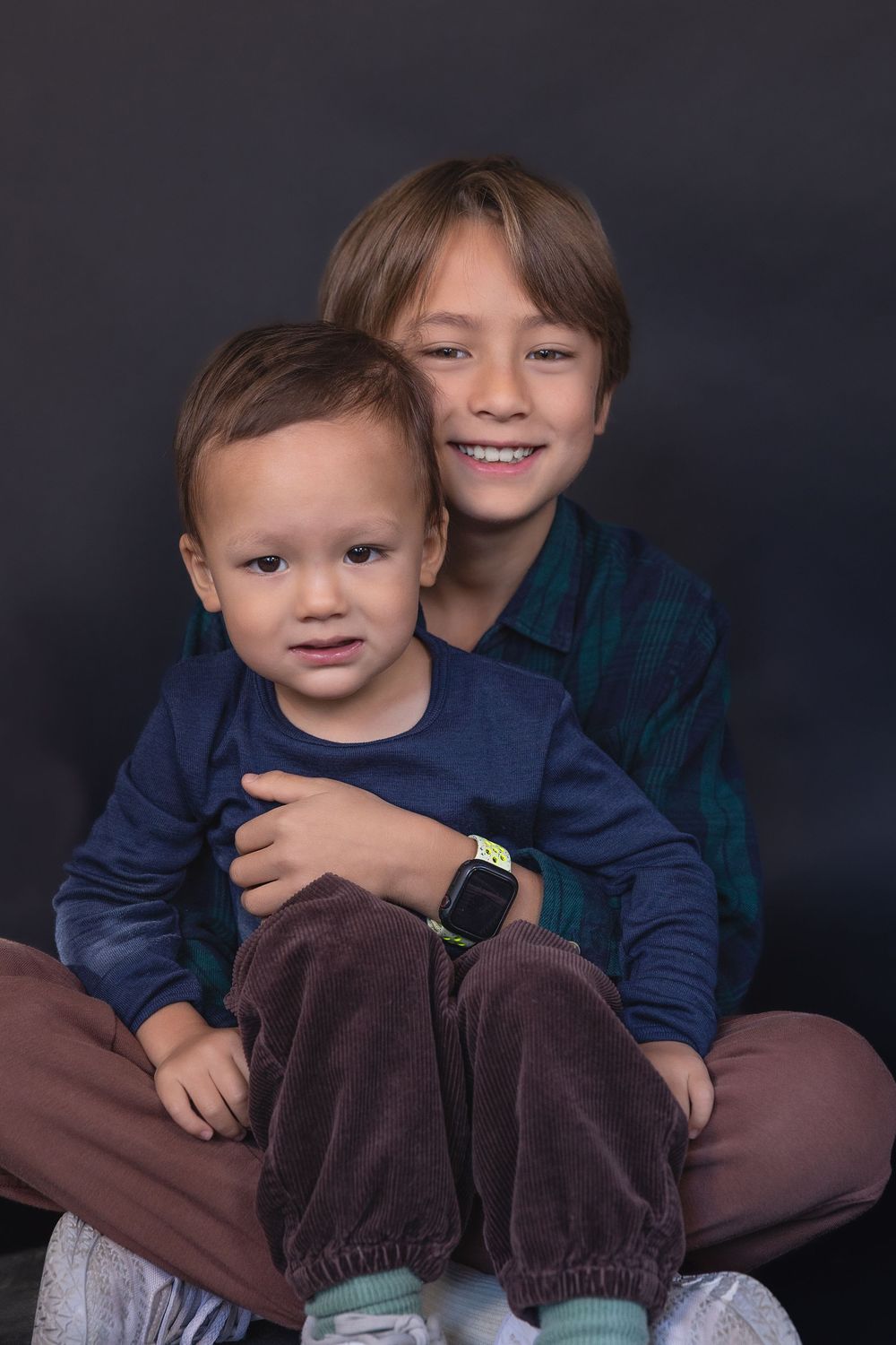 Two children in dark blue shirts sit together against a dark background for a portrait session.