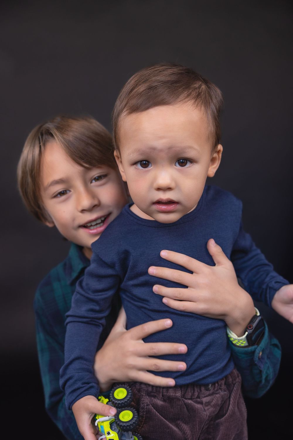 Two young children pose together against a dark background wearing blue and brown clothing.