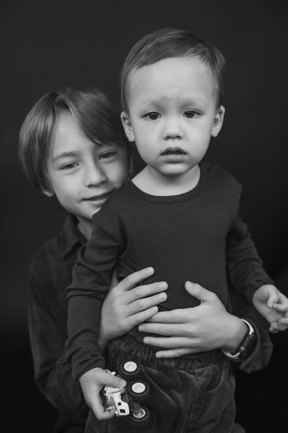 A black and white portrait of two children in dark clothing, with one holding the other from behind.