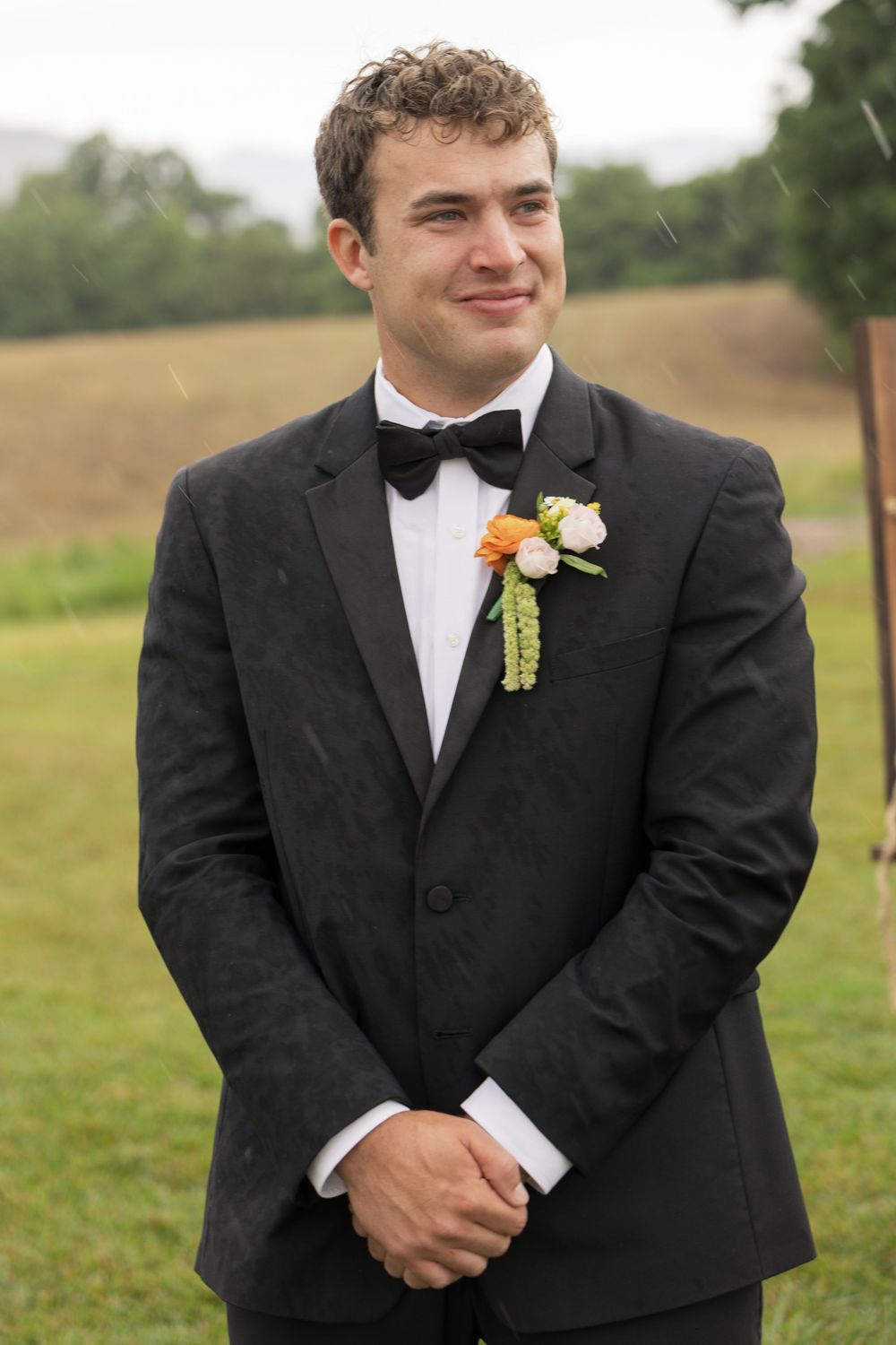 Smiling groom in black tuxedo with bow tie and floral boutonniere stands outdoors on wedding day.