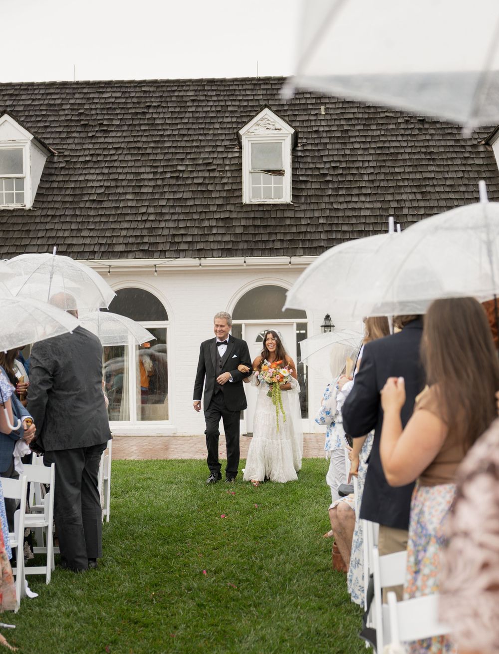 Bride and groom walk down outdoor aisle in rain as guests hold clear umbrellas at elegant white venue.