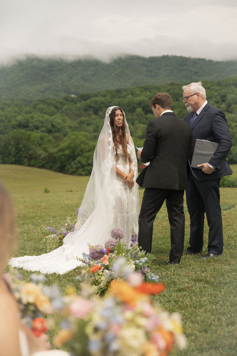 Outdoor wedding ceremony in a lush green meadow with misty mountains, bride in white gown and veil with colorful floral arrangements.