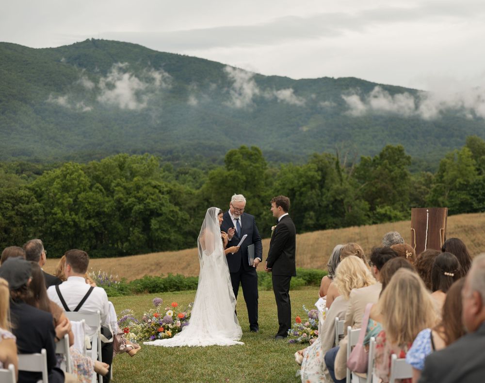 Outdoor wedding ceremony with bride and groom at altar, lush green mountains and misty clouds in background.
