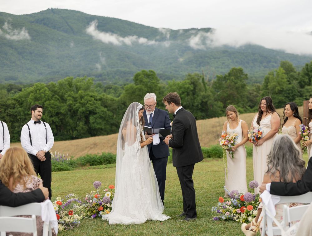 Outdoor wedding ceremony with bride and groom exchanging vows on a lush green lawn with mountain views and colorful floral arrangements.