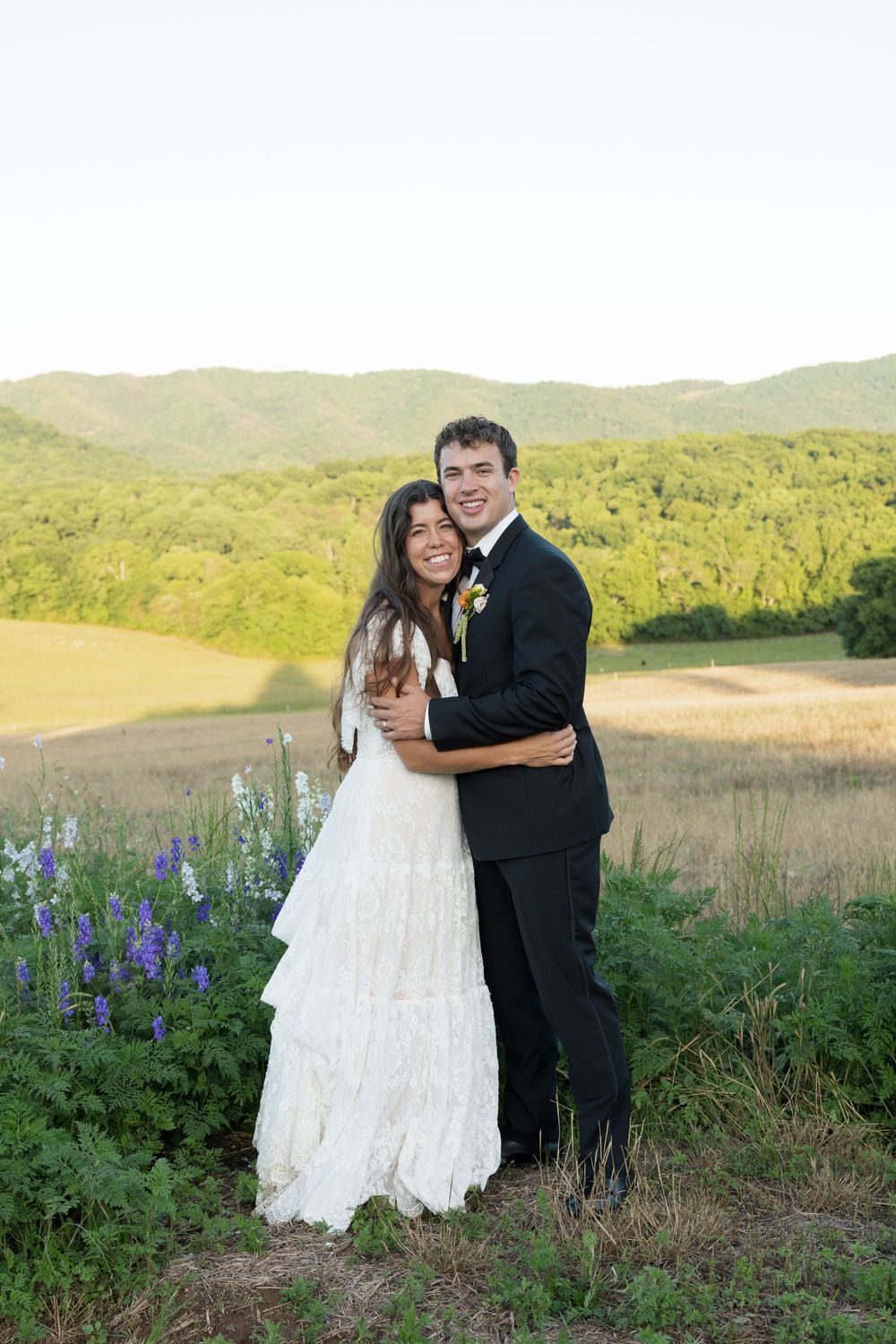 A bride and groom embrace in a scenic outdoor setting with purple wildflowers and green rolling hills in the background.