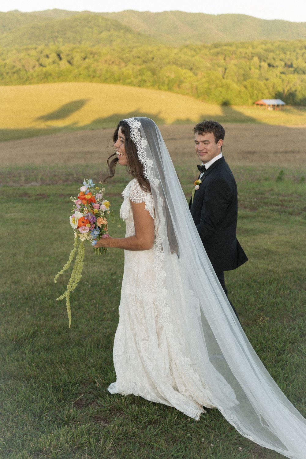 Bride in lace gown with cathedral veil holds colorful bouquet beside tuxedo-clad groom in scenic green meadow.
