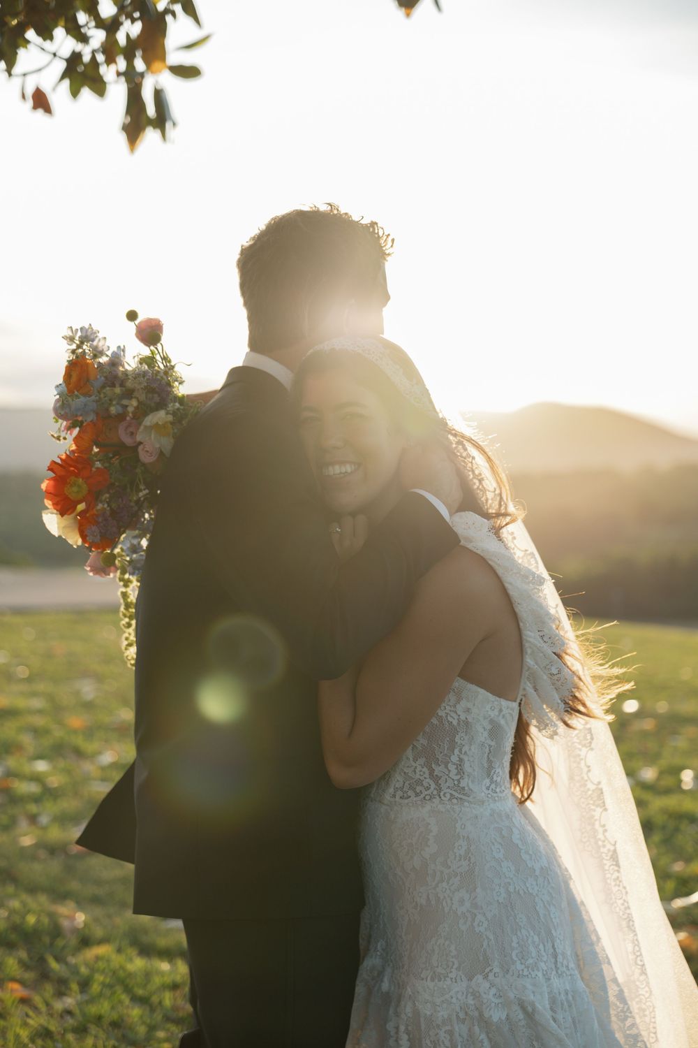 Bride in lace gown hugs groom in black suit at sunset, holding colorful bouquet with mountain backdrop.