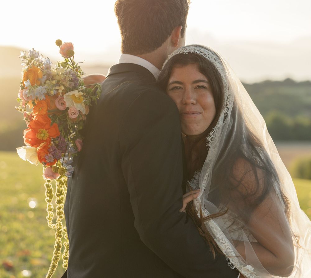 Bride in lace veil holding colorful bouquet embraces groom in black suit outdoors at golden hour.