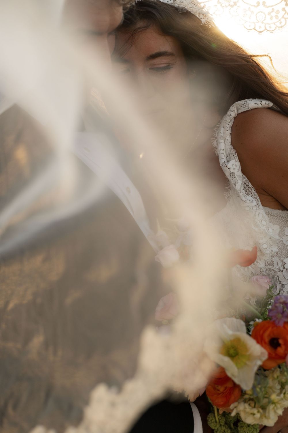 Bride in lace wedding dress holding colorful bouquet, partially obscured by flowing veil in dreamy soft light.