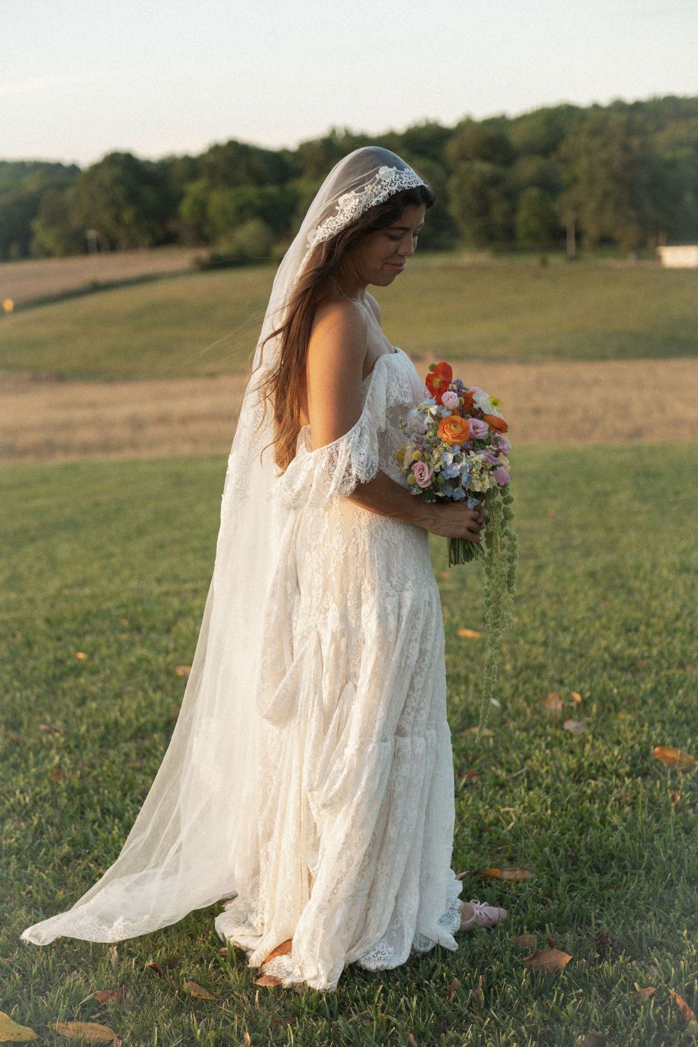 Bride in lace boho wedding dress and veil holds colorful bouquet outdoors in green field at golden hour.